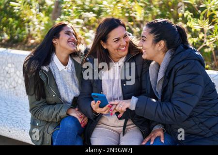 Drei weibliche Mutter und Töchter mit Smartphone sitzen auf der Bank im Park Stockfoto