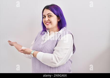 Große Frau mit lila Haaren, die auf weißem Hintergrund steht, lädt zum Eintreten ein und lächelt natürlich mit offener Hand Stockfoto