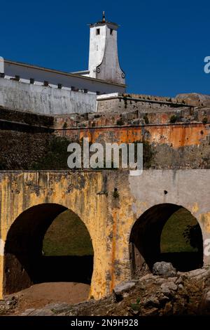 Gelb, orange und weiß: Die Burgbrücke, die zerstörten Stadtmauern und der Torre de Vigia Wachturm, dekoriert mit spiralförmigen Schriftrollen, über einem der Blöcke von Gefängniszellen, die in den 1950er Jahren im Inneren der Fortaleza oder der Festung Peniche Mitte 1500s an der Atlantikküste Portugals erbaut wurden. Politische Gegner der 1933-74 Salazar-Regierung, darunter der kommunistische Führer Álvaro Cunhal, wurden hier festgehalten, oft in Isolationszellen mit hoher Sicherheit. Stockfoto