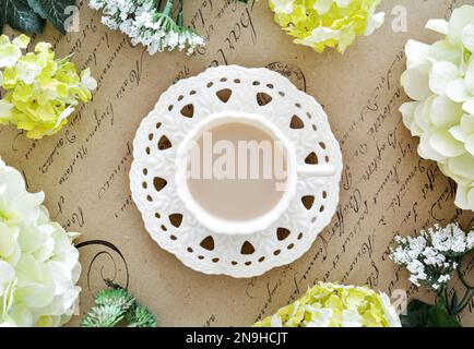 Tasse Tee mit künstlichen Blumen auf antikem Papier mit Schriftbild-Hintergrund, aufgenommen von oben in flacher Laienzusammensetzung. Stillleben im Frühling. Stockfoto