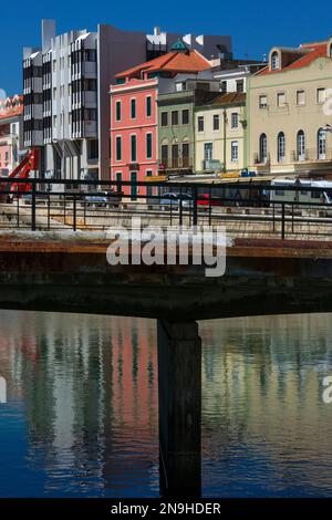 Starke zentrale Unterstützung für einen portugiesischen Fischereihafen, wobei der Stützpier einer Betonbrücke auf den ersten Blick als Stütze für die farbenfrohe Uferpromenade von Peniche im Stadtteil Leiria im Zentrum Portugals mit Bars, Restaurants, Geschäften und Wohnblocks erscheint. Unterhalb der Brücke teilt der Pier eine schimmernde Reflexion der sonnigen Sommerszene. Stockfoto