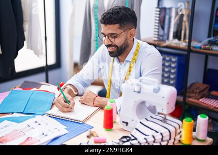 Ein junger arabischer Schneider, der selbstbewusst in einem Schneiderladen auf ein Notebook lächelt Stockfoto