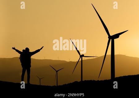 Silhouetten eines Reisenden mit Rucksack und Windturbinen bei Sonnenuntergang Stockfoto