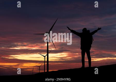 Silhouetten eines Reisenden mit Rucksack und Windturbinen bei Sonnenuntergang Stockfoto