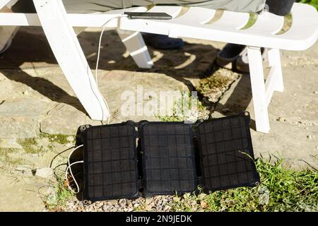 Die Solarbatterie liegt auf dem Boden in der Nähe des Plastikstuhls, auf dem das Mobiltelefon aufgeladen wird. Konzept alternativer Energiequellen. Stockfoto