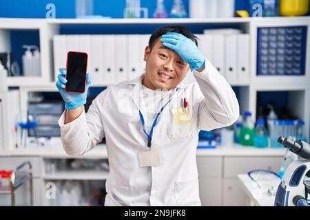 Ein junger chinesischer Mann, der im Labor für Wissenschaftler arbeitet, hält sein Smartphone gestresst und frustriert von Hand an Kopf, überrascht und wütend Stockfoto