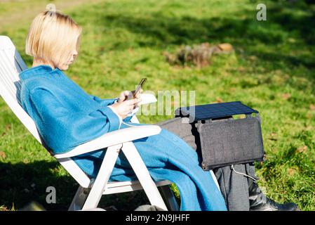 Eine Frau in einem Mantel mit einem Mobiltelefon in der Hand und einer tragbaren Solarbatterie sitzt im Garten. Ökologisches Alternativenergiekonzept. Stockfoto
