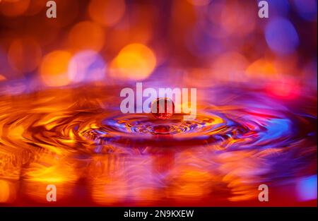 Ein transparenter Tropfen mit rot-gelbem Hintergrund fällt ins Wasser. Abstrakter, farbenfroher Hintergrund Stockfoto