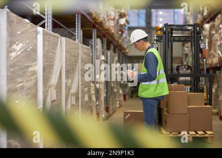 Seitliches Porträt eines jungen männlichen Arbeiters, der im Lagerhaus eine Bestandsaufnahme durchführt und auf das Klemmbrett schreibt Stockfoto