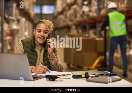 Porträt einer lächelnden Arbeitnehmerin, die im Lager ein Notebook benutzt und telefonisch das Vertriebsgeschäft leitet, Kopierraum Stockfoto