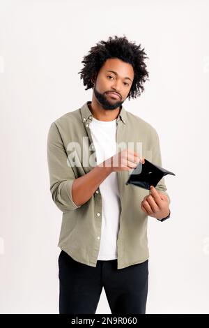 Deprimierter afroamerikanischer junger Mann zeigt leere Brieftasche isoliert auf weißem Hintergrund, aufgebrachter, lockiger Kerl hat Geldmangel, schwierige finanzielle Lage aufgrund der Wirtschaftskrise Stockfoto