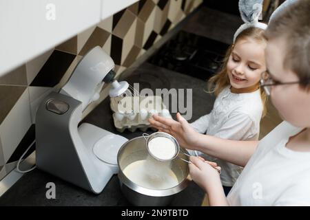 Bruder, der Mehl siebt, Teig für Kekse zubereitet, Brötchen gekreuzt. Schwester in Hasenohren, die sich den Prozess anschaute. Ostern, Familienunterstützungskonzept Stockfoto