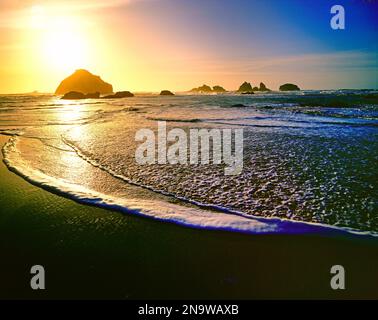 Helle untergehende Sonne, die unter dem Horizont über dem Wasser mit Felsformationen entlang der Küste Oregons, Oregon, Vereinigte Staaten von Amerika, untergeht Stockfoto