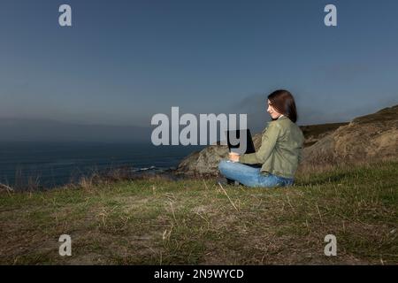 Frau, die Freiberuflerin, die auf einer hohen Klippe über dem Meer sitzt und aus der Ferne arbeitet Stockfoto