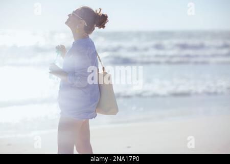 Lächelnde elegante 40-jährige Frau mit weißem Strohbeutel und Getränk am Strand. Stockfoto