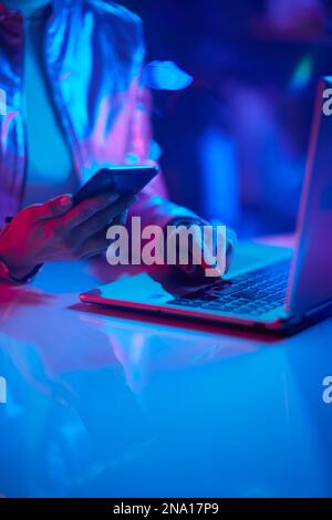 Neon-Metaverse-futuristisches Konzept. Nahaufnahme einer Frau mit Laptop, die im Büro Smartphone-Anwendungen verwendet. Stockfoto