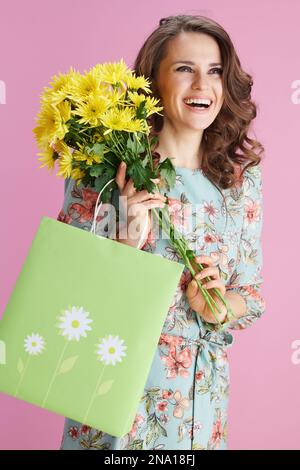 Lächelnde elegante Frau mittleren Alters in Blumenkleid mit gelben Chrysanthemen und grüner Einkaufstasche isoliert auf pinkfarbenem Hintergrund. Stockfoto