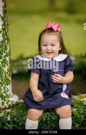 Ein süßes kleines, zwei Jahre altes Mädchen, das draußen in einem marineblauen Kleid sitzt Stockfoto