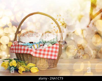 Korb mit köstlichen Osterkuchen, bemalten Eiern und Blumen auf einem Holztisch. Platz für Text Stockfoto