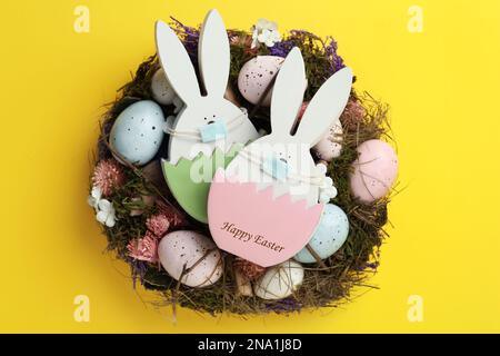 Holzhasen mit Schutzmasken, bemalten Eiern und Osterkranz auf gelbem Hintergrund, Draufsicht. Urlaub während der COVID-19-Quarantäne Stockfoto