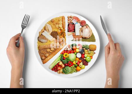 Frau hält Besteck in der Nähe des Tellers mit verschiedenen Produkten auf weißem Hintergrund, Draufsicht. Ausgewogenes Essen Stockfoto