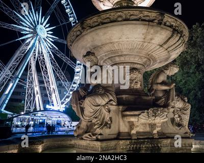 Ein beleuchtetes Fährrad und Gebäude mit einer geschnitzten Steinstatue im Vordergrund bei Nacht; Budapest, Budapest, Ungarn Stockfoto