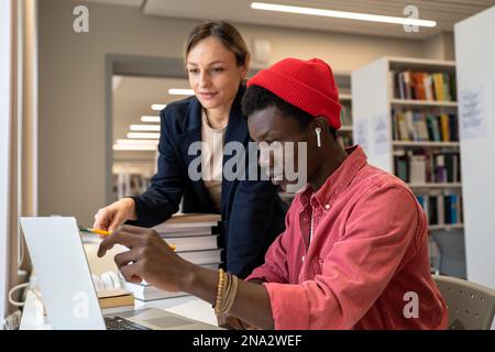 Eine Privatlehrerin, die einem verwirrten, fokussierten schwarzen Schülertyp bei der Lösung von Aufgaben auf einem Laptop in der Bibliothek hilft. Stockfoto