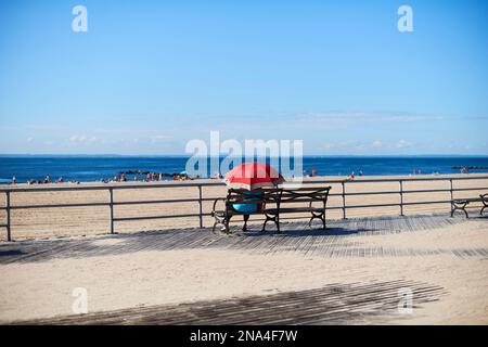 Frau sitzt mit Schirm auf der Promenade von Coney Island mit Blick auf den Atlantischen Ozean; New York City, New York, Vereinigte Staaten von Amerika Stockfoto