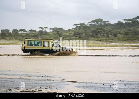 Das Safari-Fahrzeug spritzt durch die überflutete Schlucht im Ndutu-Gebiet des Ngorongoro-Krater-Schutzgebiets auf den Serengeti-Ebenen in Tansania. Stockfoto