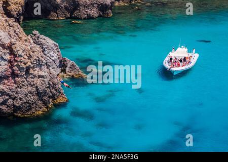 Touristen auf einem Ausflugsboot am Tsigado Beach, mit einem jungen Mann, der von einer Klippe springt; Milos Island, Kykladen, Griechenland Stockfoto