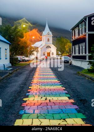 Der farbenfrohe Pfad lag mitten in einer Asphaltstraße, der zu einer weißen Kirche führte; Seydisfjordur, Eastern Region, Island Stockfoto