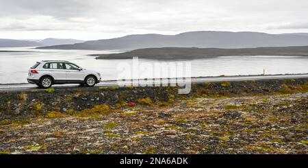 Ein Fahrzeug, das auf einer Straße am Ufer des Wassers geparkt ist, mit Blick auf den Fjord und die Silhouette der Hügel unter einem bedeckten Himmel; Sudavik, Westfjorde, Island Stockfoto