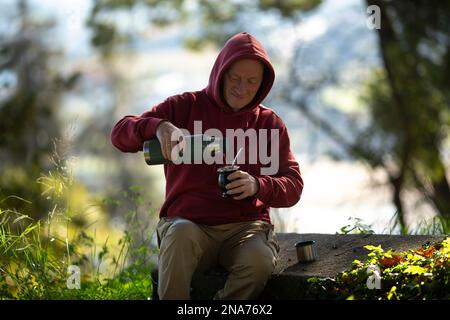 Ein Mann schüttet Mate aus einer Thermoskanne in eine Tasse im Park. Stockfoto