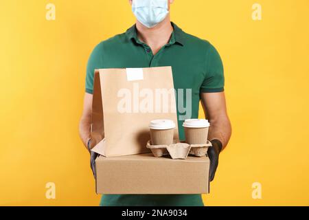 Kurier in der medizinischen Maske hält Pakete mit Essen und Getränken zum Mitnehmen auf gelbem Hintergrund, Nahaufnahme. Zustellung während der Quarantäne wegen Covid- Stockfoto