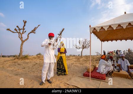 Rajput-Mann spielt Ravanahatha und Sängerin tritt beim Festival mit Dorfältesten auf, die in der Wüste Thar in Rajasthan, Indien, eine Wasserpfeife rauchen Stockfoto