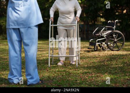 Ältere asiatische Seniorin praktizieren mit einem Aluminiumwandler zur Sicherheit mit einer Krankenschwester. Pflegeheim Krankenhaus-Garten-Konzept Stockfoto