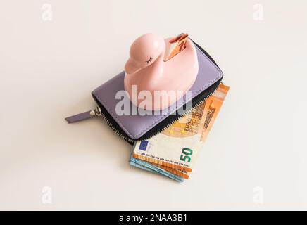 Rosafarbene Keramikmoneybox auf der lila Lederbörse mit Euro-Banknoten auf hellem Hintergrund. Stockfoto