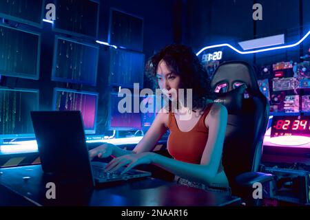 Weibliche asiatische Hacker in einem Versteck mit dunkler Umgebung und mehreren Displays und Kabeln, versuchen in Datenserver einzubrechen und das System mit einem Virus zu infizieren. Stockfoto