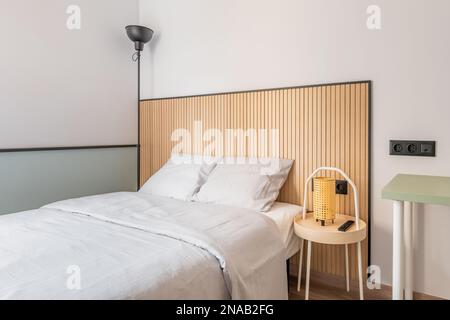 Teil eines Schlafzimmers mit einem Doppelbett an einer weißen Wand mit Designer-Holzelementen. Stehleuchte mit schwarzem Metallschirm in der Ecke. Auf der Stockfoto