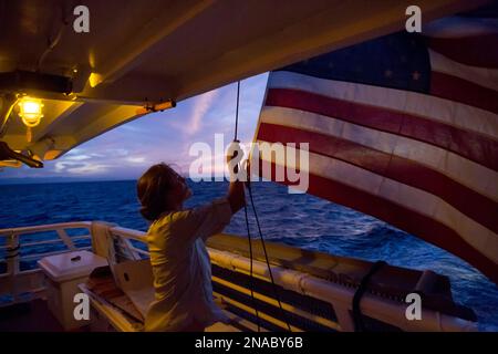 Inmitten eines farbenfrohen Morgenhimmels in der Nähe von Panama hebt ein Deckblatt an Bord eines Expeditionsschiffs bei Sonnenaufgang im Coiba-Nationalpark, Panama, die amerikanische Flagge Stockfoto