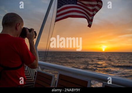 Inmitten eines farbenfrohen Morgenhimmels in der Nähe von Panama fotografiert ein Passagier an Bord eines Expeditionsschiffs den Sonnenaufgang, während eine amerikanische Flagge im Wind weht Stockfoto