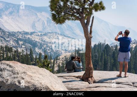 Am Olmstead Point trübt der Rauch eines Waldfeuers den Blick auf den Yosemite National Park, Kalifornien, während ein Paar umarmt und ein Mann fotografiert Stockfoto