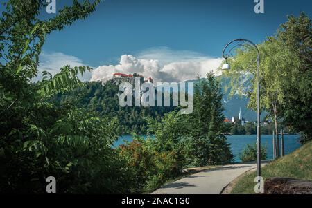 Spaziergang am Bleder See zwischen Bäumen und Straßenlaternen mit einer Burg Bled auf dem Felsen und der Kirche Zupnijska cerkev svetega Martina Stockfoto