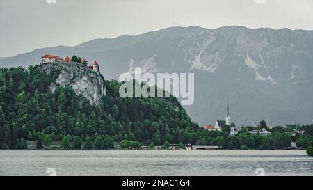 Bled See mit einem Bled Schloss auf dem Felsen und Kirche Zupnijska cerkev svetega Martina - Kirche St. Martina, Bled, grauer Himmelshintergrund Stockfoto