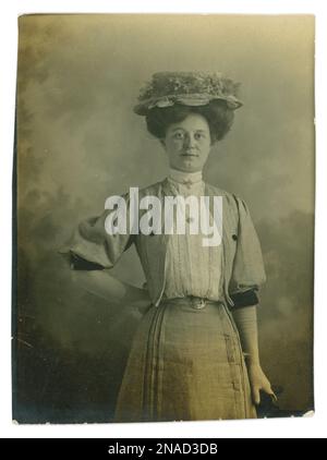 Originales edwardianisches Foto von einer hübschen Arbeiterfrau namens Amy. Edwardianerin. Edwardianerin. Sie war vielleicht die Dienstmädchen einer Dame. Sie trägt einen Strohhut / Bootsboot mit Blumen um die Krempe, große Frisur, Wishbone-Stil Brosche, weiße Handschuhe, Gürtel um eine sehr schleimige Taille, feine Plissee-Sommerbluse aus Baumwolle mit kleinen Punkten, mit hohem Ausschnitt, Bolero-Jacke mit Ärmeldetails - durchgehende Ärmel bis Ellbogen und passender Rock. Vom 5. Juni 1907. Stockfoto