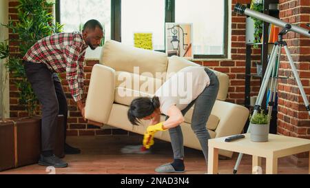 Ein junges Paar wäscht den Holzboden mit Mopp, der Ehemann hilft der Frau, unter der Couch zu putzen. Mann und Frau machen Frühjahrsputz im Wohnzimmer, mit Allzweckreiniger und Waschlösung. Stockfoto