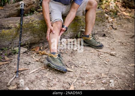 Das abgeschnittene Bild eines männlichen Trekkers sitzt auf einem Holzstamm und massiert sein Bein, leidet an Knieschmerzen, Beinkrämpfen, verletzt während des Weges. Stockfoto