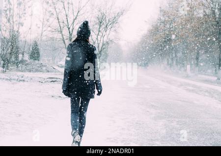 Eine Frau in Schwarz läuft durch einen Schneesturm auf einer verschneiten Straße Stockfoto