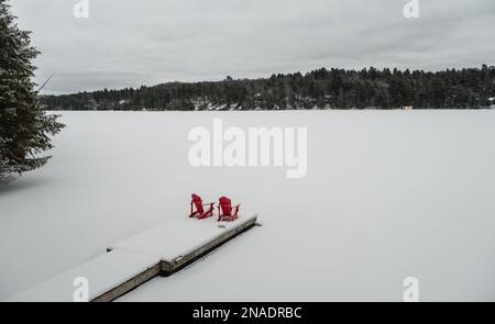 Rote Adirondack-Stühle am Ende des verschneiten Docks auf einem See im Winter. Stockfoto