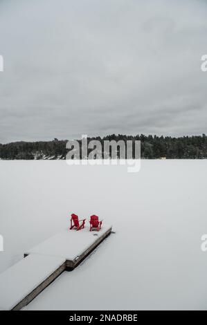 Rote Adirondack-Stühle am Ende des verschneiten Docks auf einem See im Winter. Stockfoto